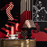Close-up of red and black checkered wallpaper, brass cabinet, and checkered lampshade with black fringe.