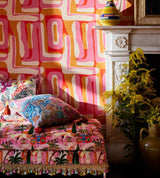 A vibrant, bohemian bedroom with a bold pink and orange abstract-patterned wallpaper. The eclectic decor features a mix of floral cushions, a fringed daybed, and a classic white fireplace adorned with yellow flowers