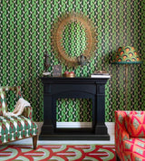 A maximalist living space featuring green geometric wallpaper, a black fireplace, a sunburst mirror, patterned armchairs, and vibrant decor accents