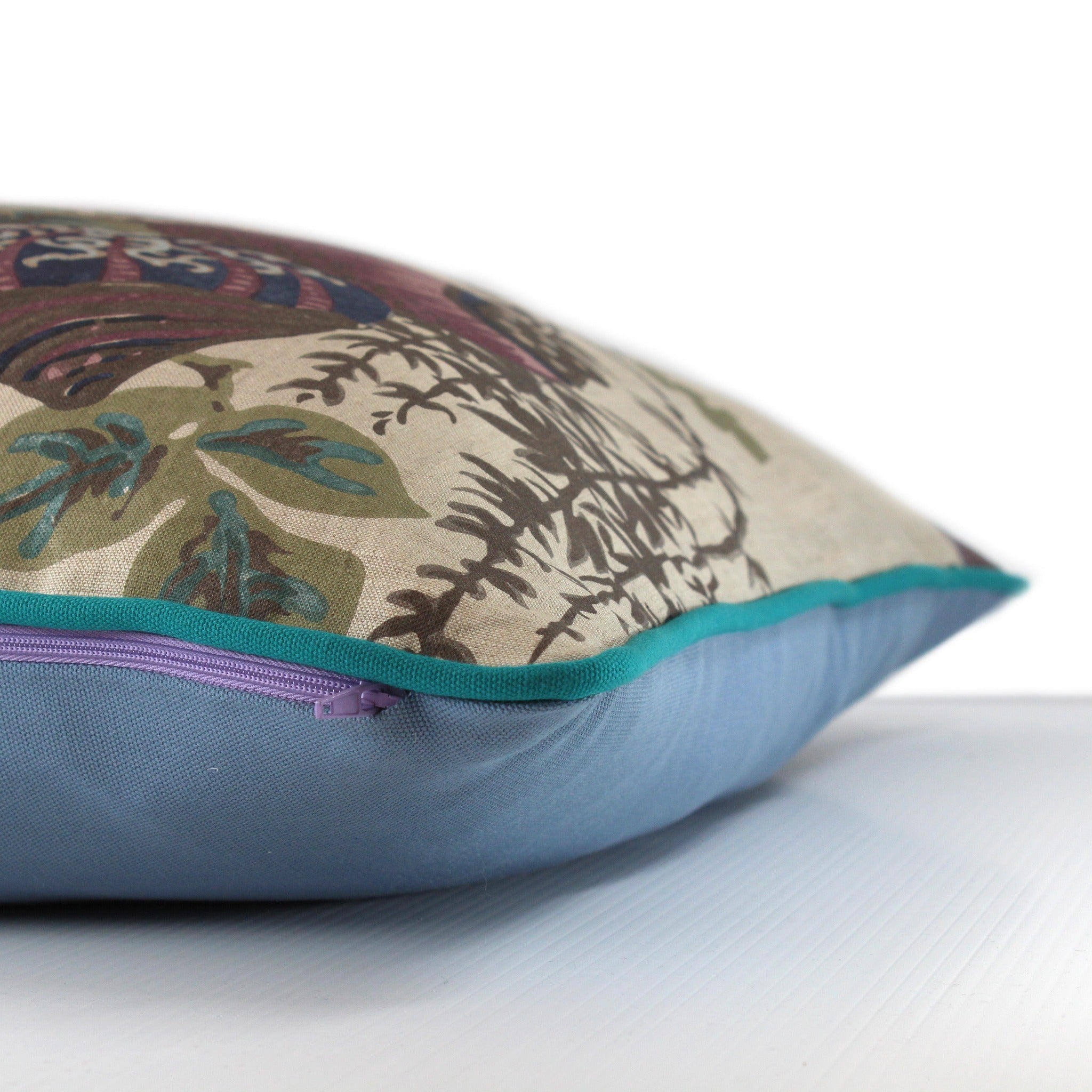 Side view of a floral cushion, natural linen base cloth with dusty purple , navy, powder blue and verdigris coloured large scale flowers and turquoise piping