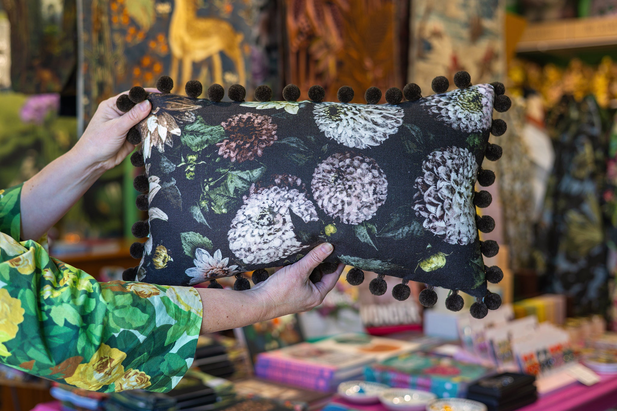 A person holds a rectangular decorative pillow with a black background, a large white and green floral print, and dark pom-pom trim along the edges in a shop setting.