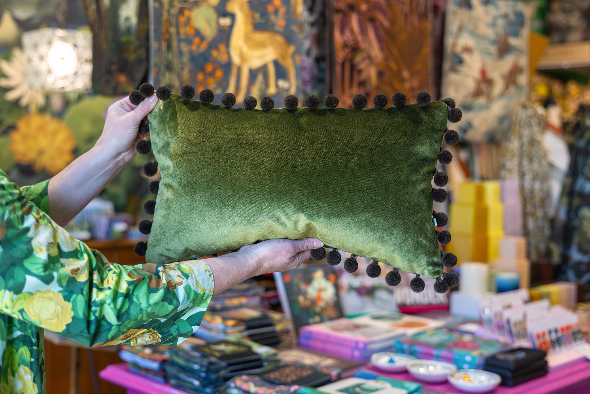 A person holds up an olive green velvet rectangular pillow with dark brown pom-pom trim, set against a colorful, busy artisan shop background.
