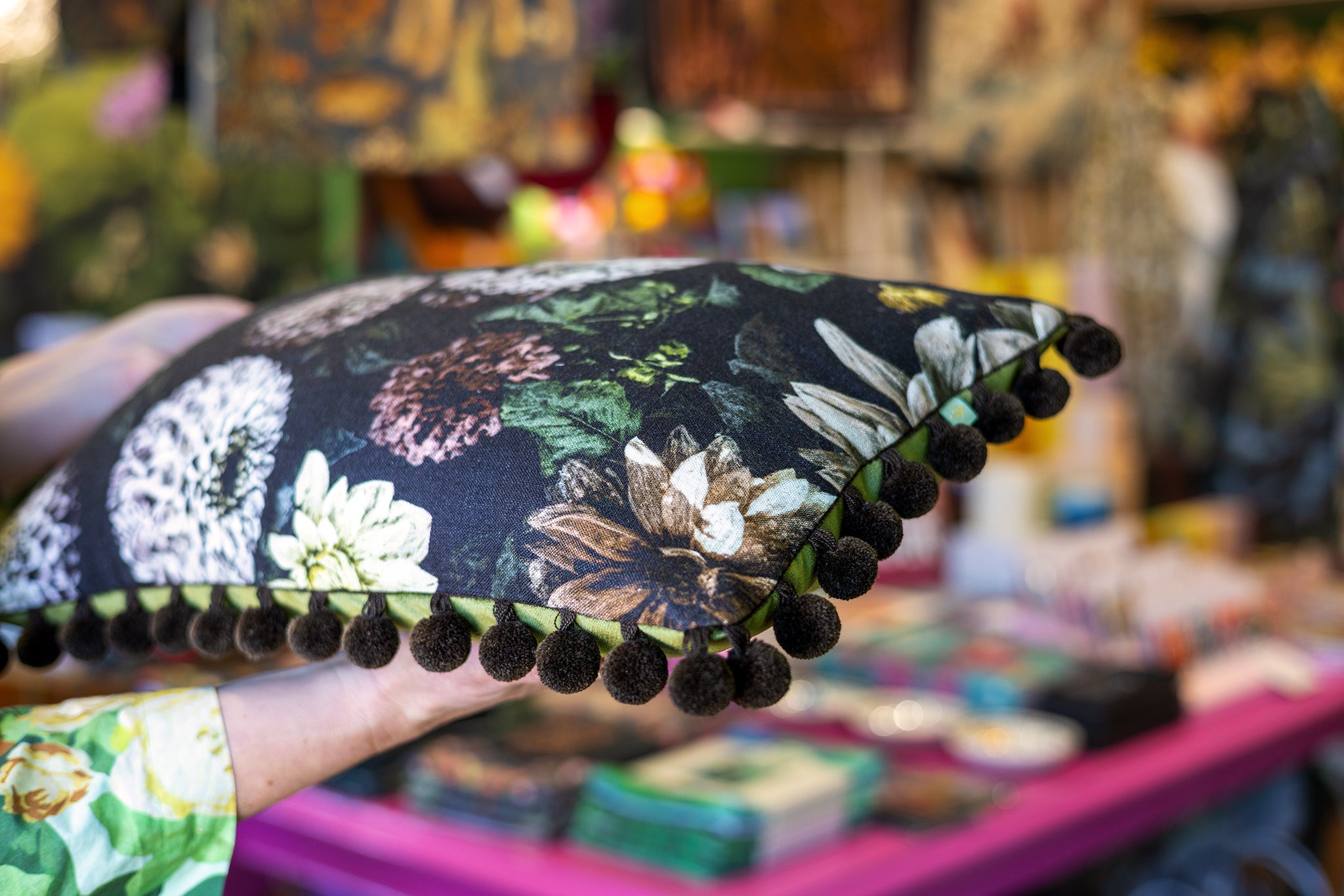 A hand holds a square decorative pillow featuring a black background with a multicolor floral print and dark pom-pom trim, set against a blurred market stall background.