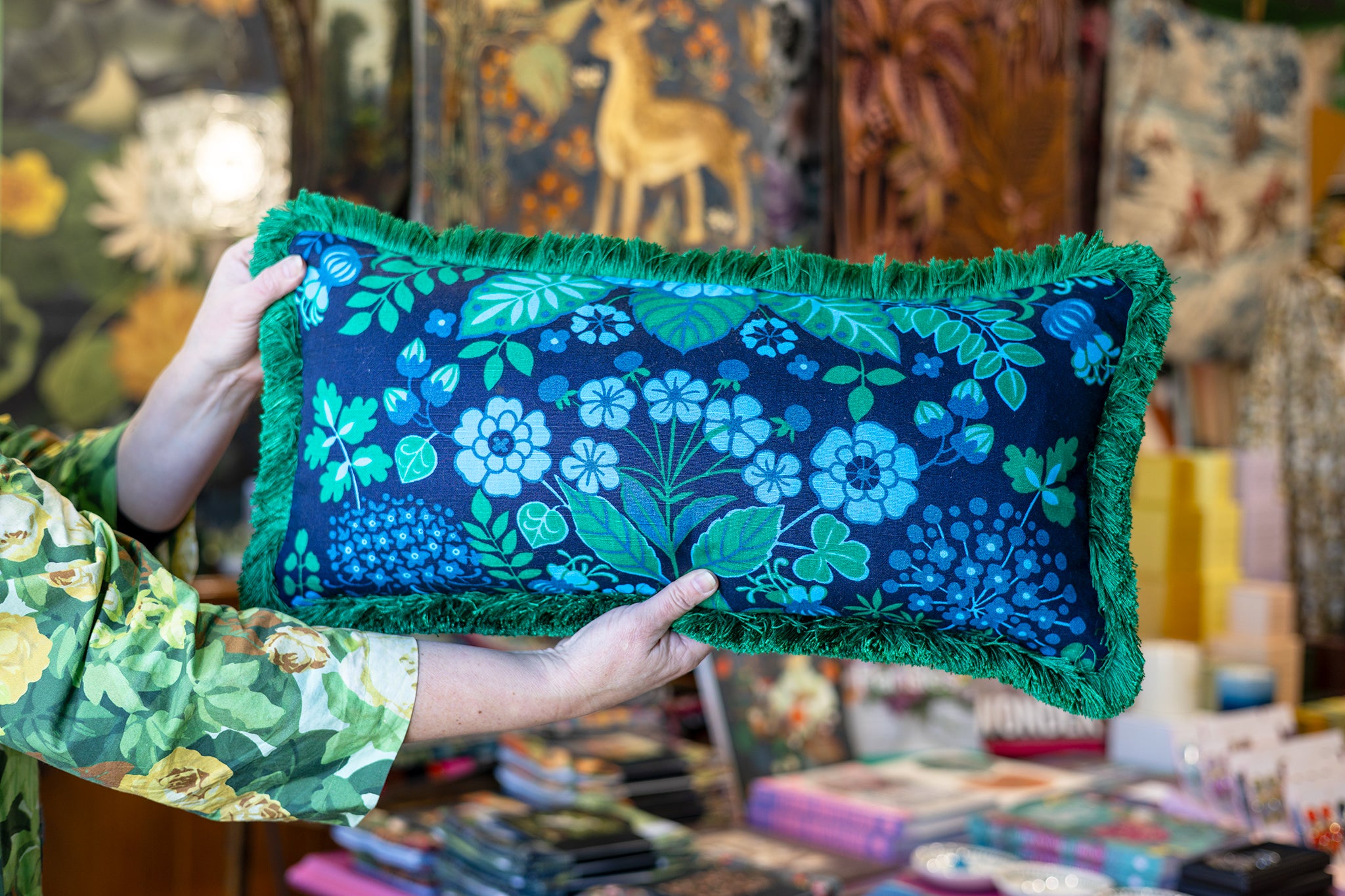 A person holds a rectangular decorative pillow with a dark navy blue background, a vibrant green and blue floral pattern, and green fringe trim, set against a busy, colorful shop background.