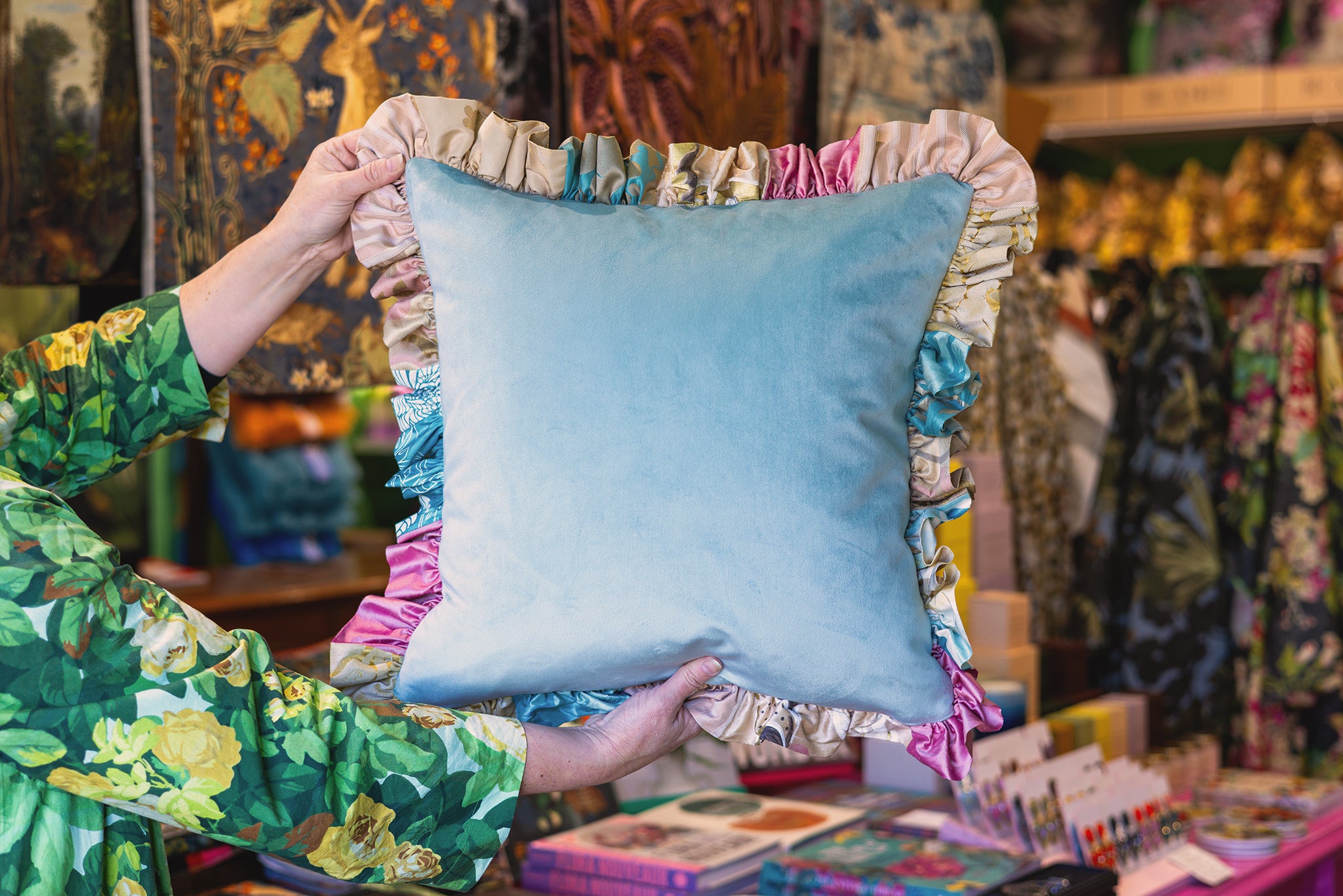 A person in a green floral dress holds up a square light blue velvet pillow with a ruffled border in pastel colors, set against a busy, colorful shop background.