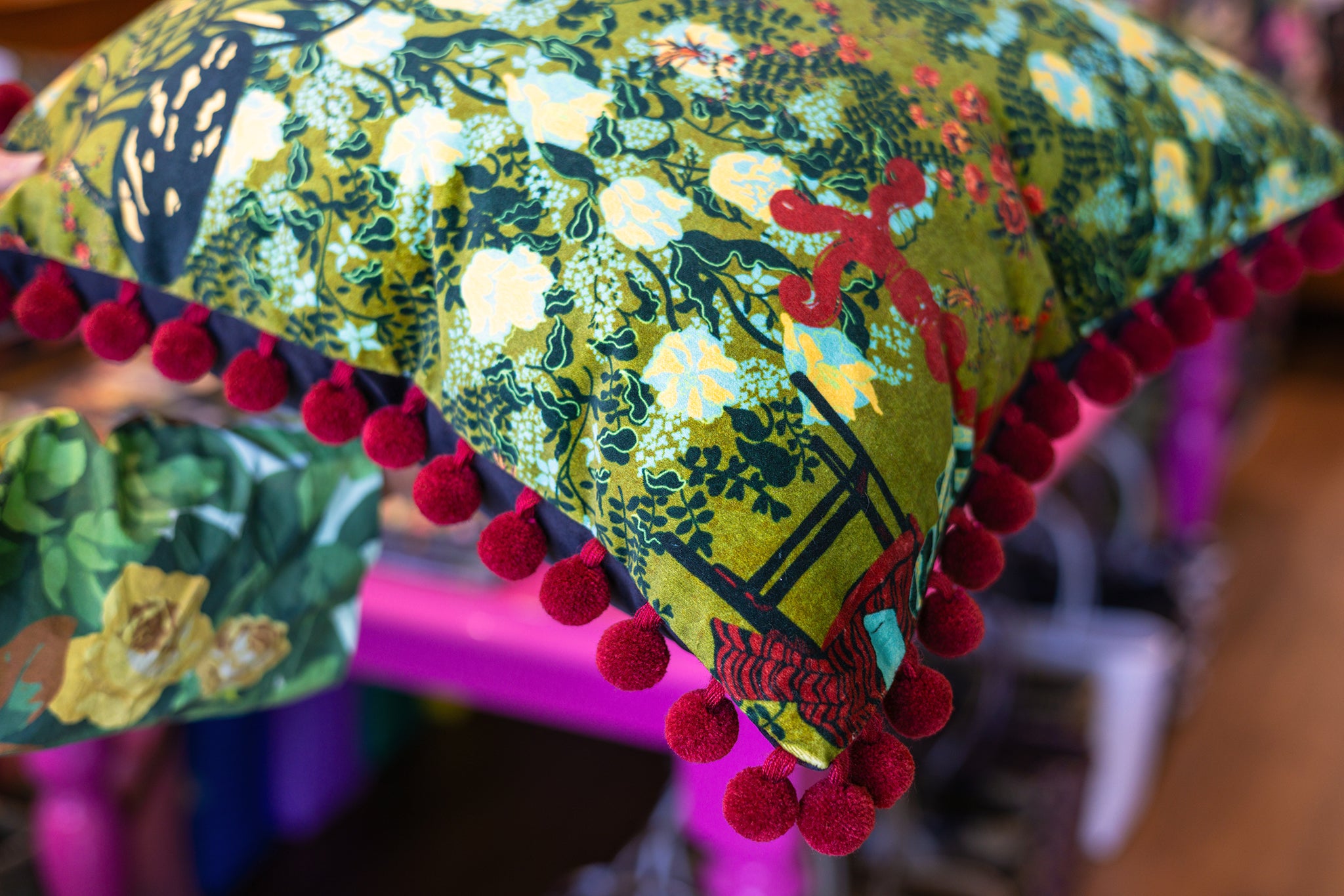 A close-up of a decorative throw pillow featuring a green and yellow botanical pattern with dark green leaves, white and yellow flowers, and a red abstract design. The pillow is finished with a prominent row of red pom-poms along the edge.