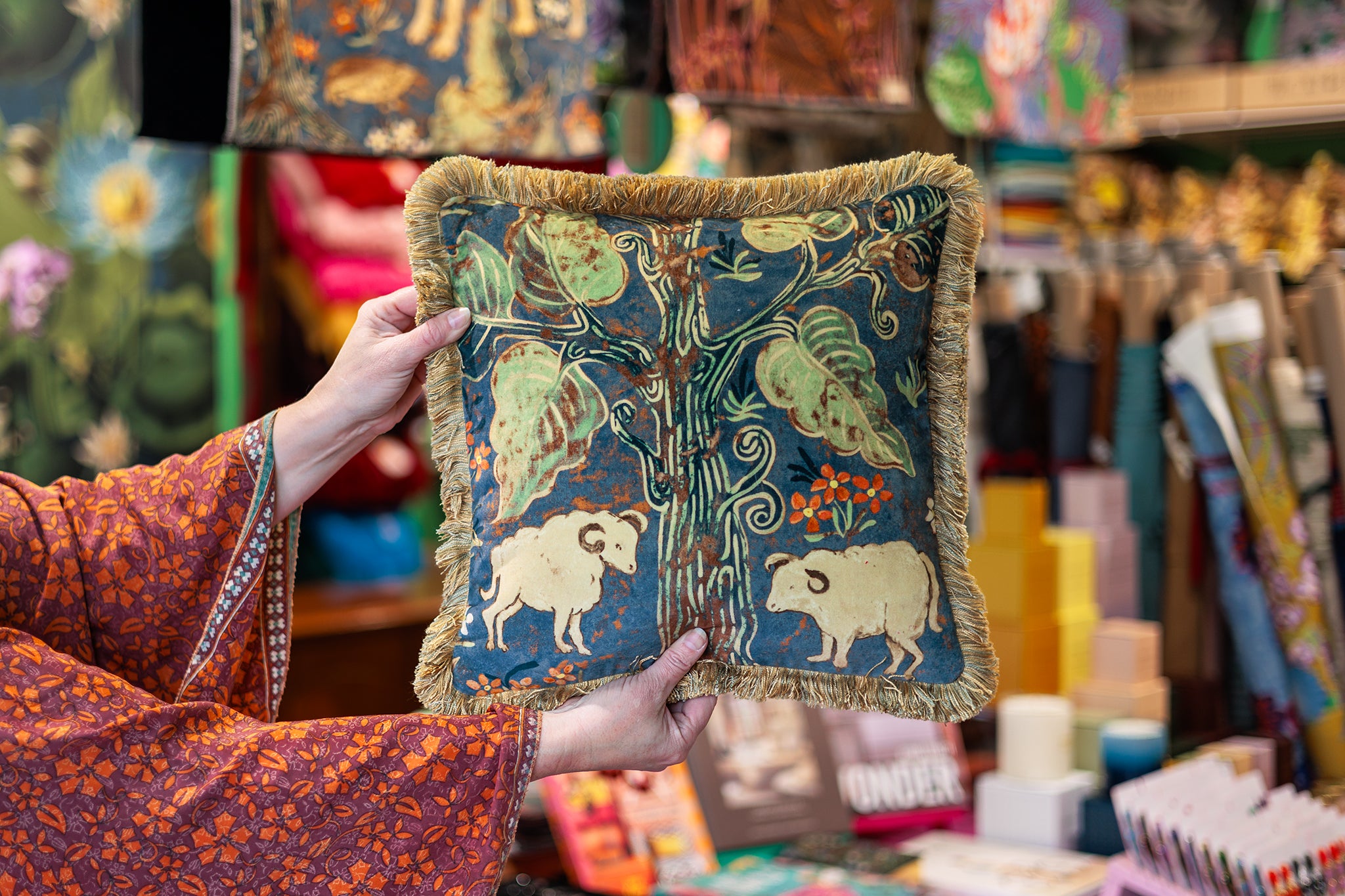 A person holds a square decorative throw pillow in a colorful shop. The pillow features a dark teal-blue background with a vintage-style illustration of a tree, leaves, and two white sheep, finished with a gold fringe border.