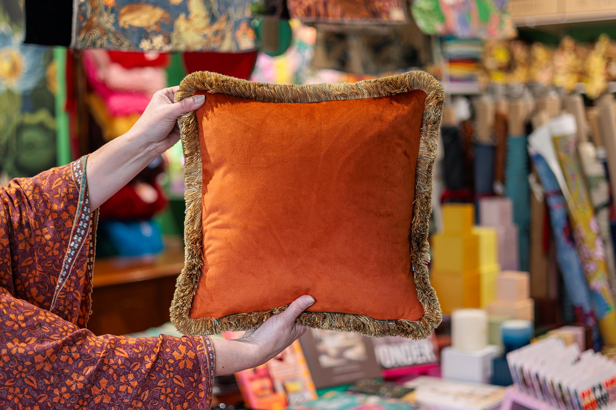 A person holds up a square orange velvet throw pillow with a textured, fringed border in a colorful, busy shop setting.