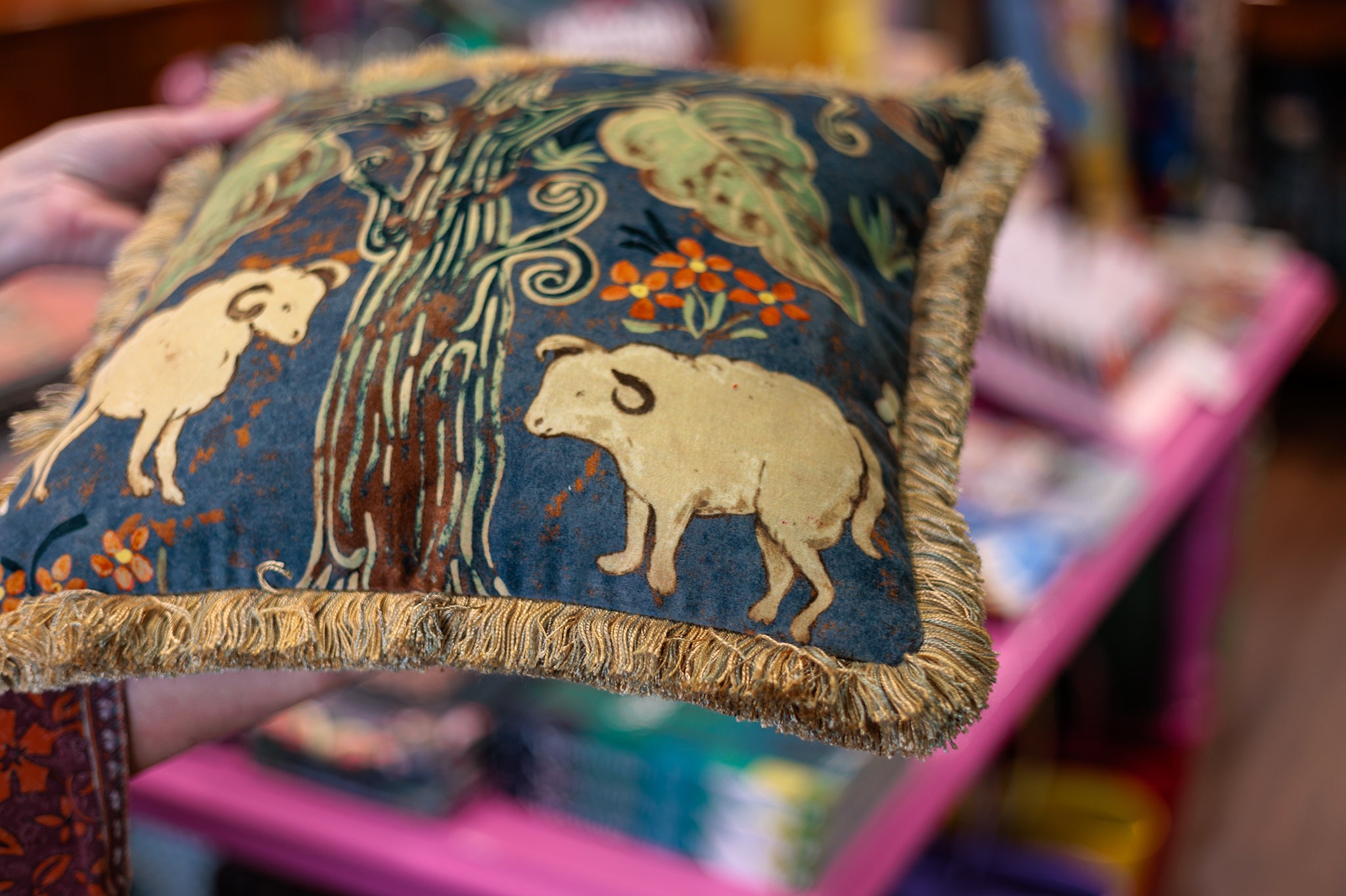 A close-up of a hand holding a square decorative throw pillow with a dark blue background, featuring cream-colored sheep, a tree, and orange floral motifs, finished with a fringed edge.