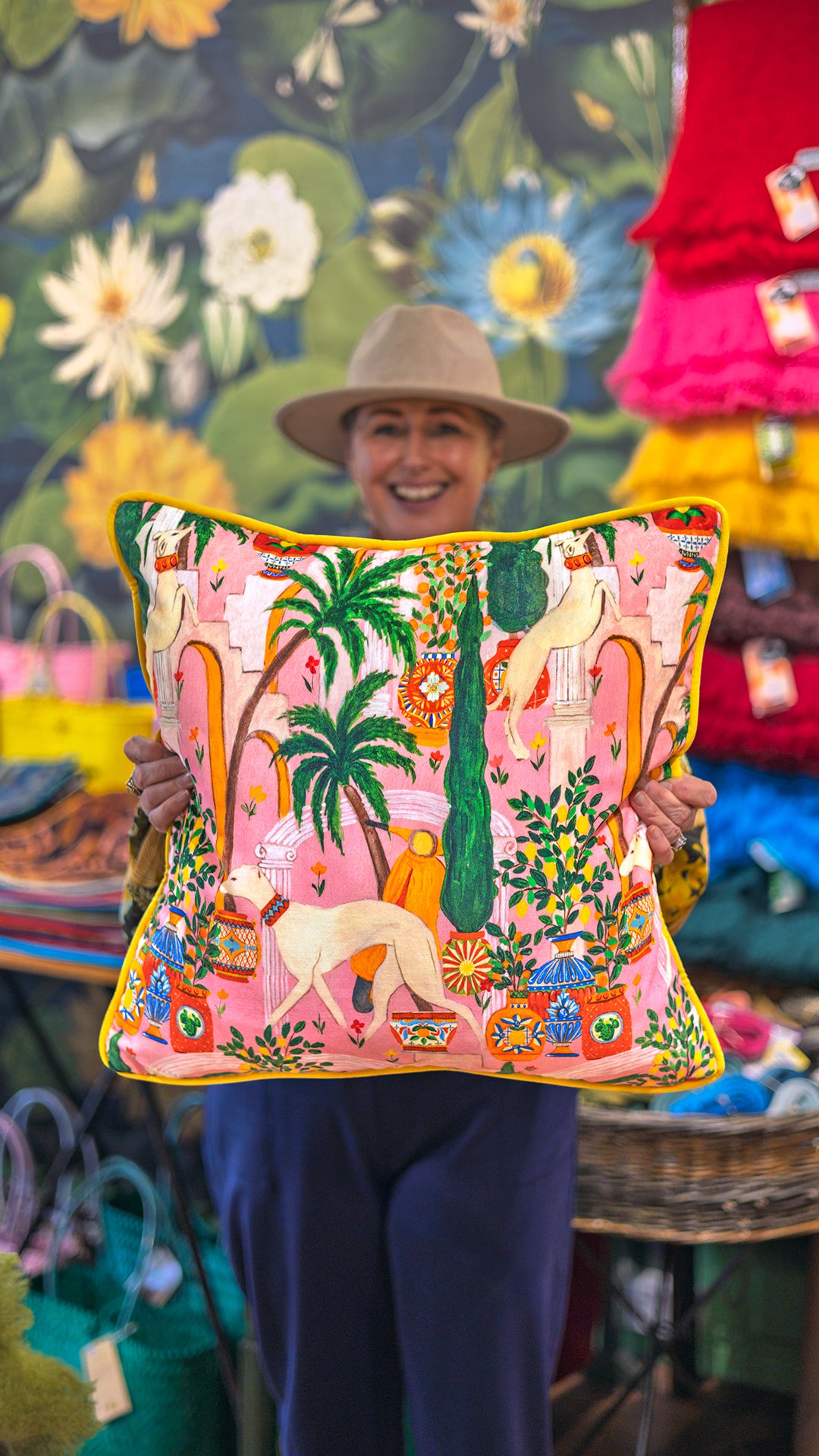 A smiling woman in a beige hat and navy pants holds up a square decorative throw pillow at a boutique. The pillow features a vibrant pink background with a whimsical illustrated print of palm trees, birds, and animals, bordered by a bright yellow trim.