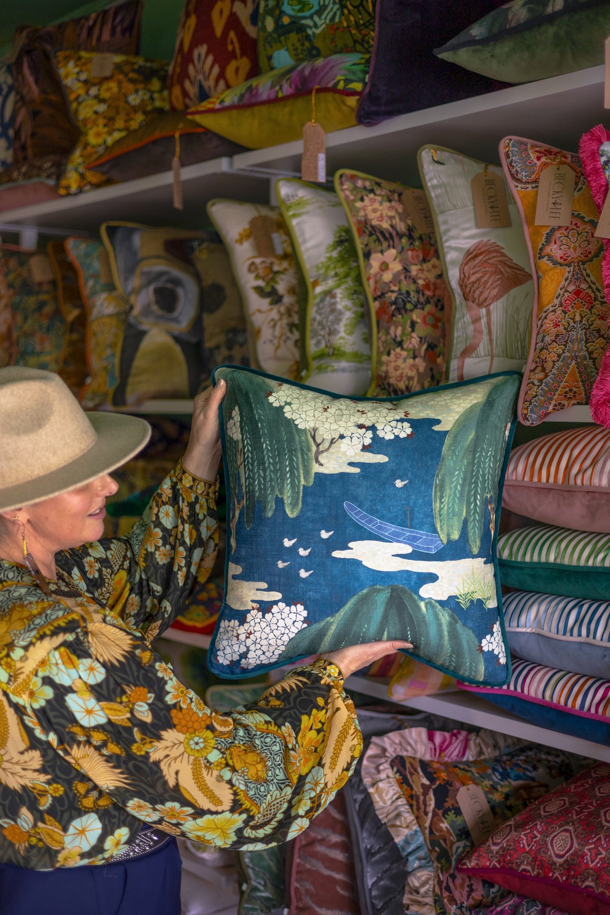 A woman in a patterned blouse and wide-brim hat holds up a teal blue decorative throw pillow with a landscape print, surrounded by shelves of other colorful pillows in a shop.