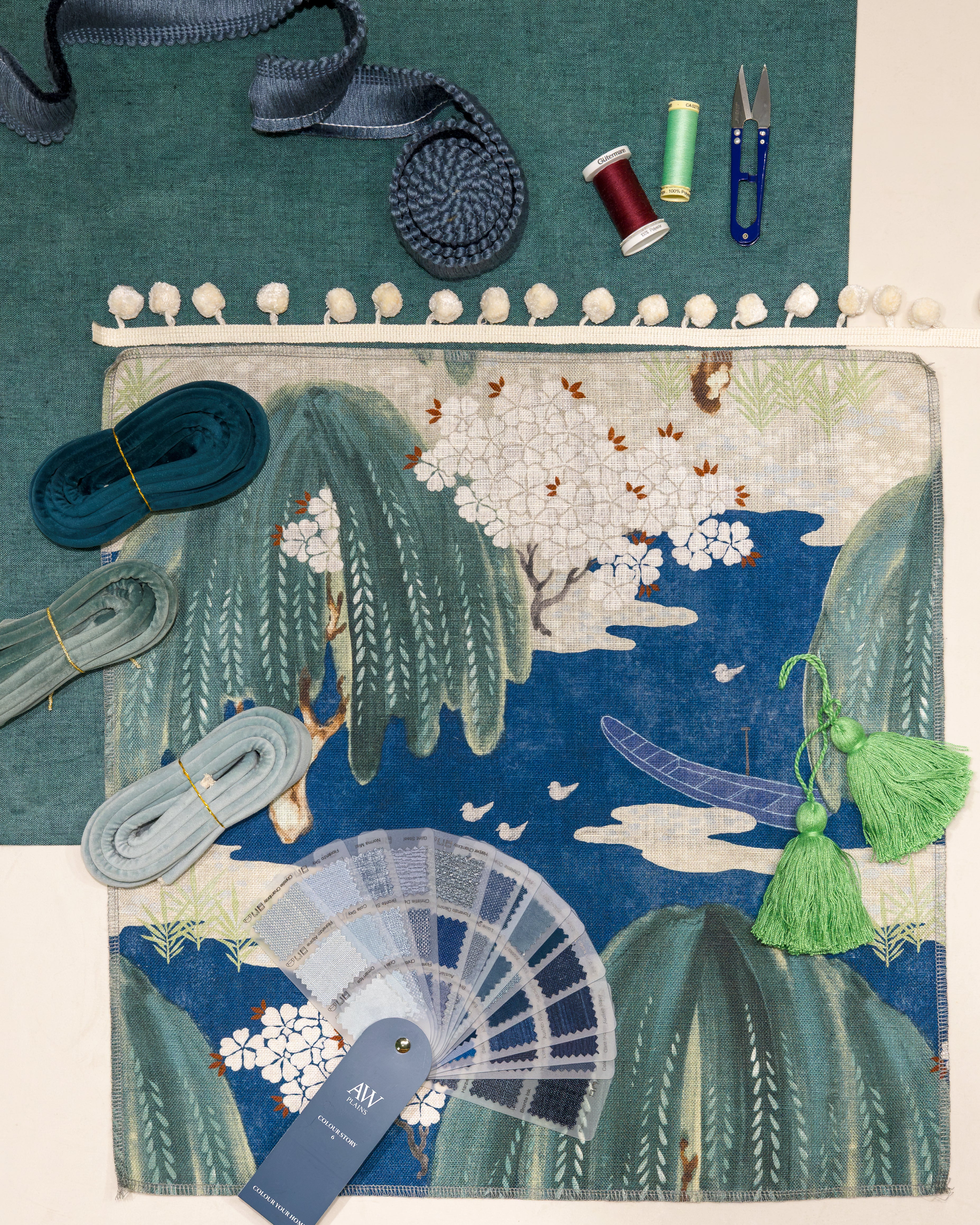 A styled flat-lay of a Japanese-style textile design featuring a blue and green landscape with white cherry blossoms, surrounded by crafting supplies including spools of thread, ribbon, scissors, and a fabric swatch fan.
