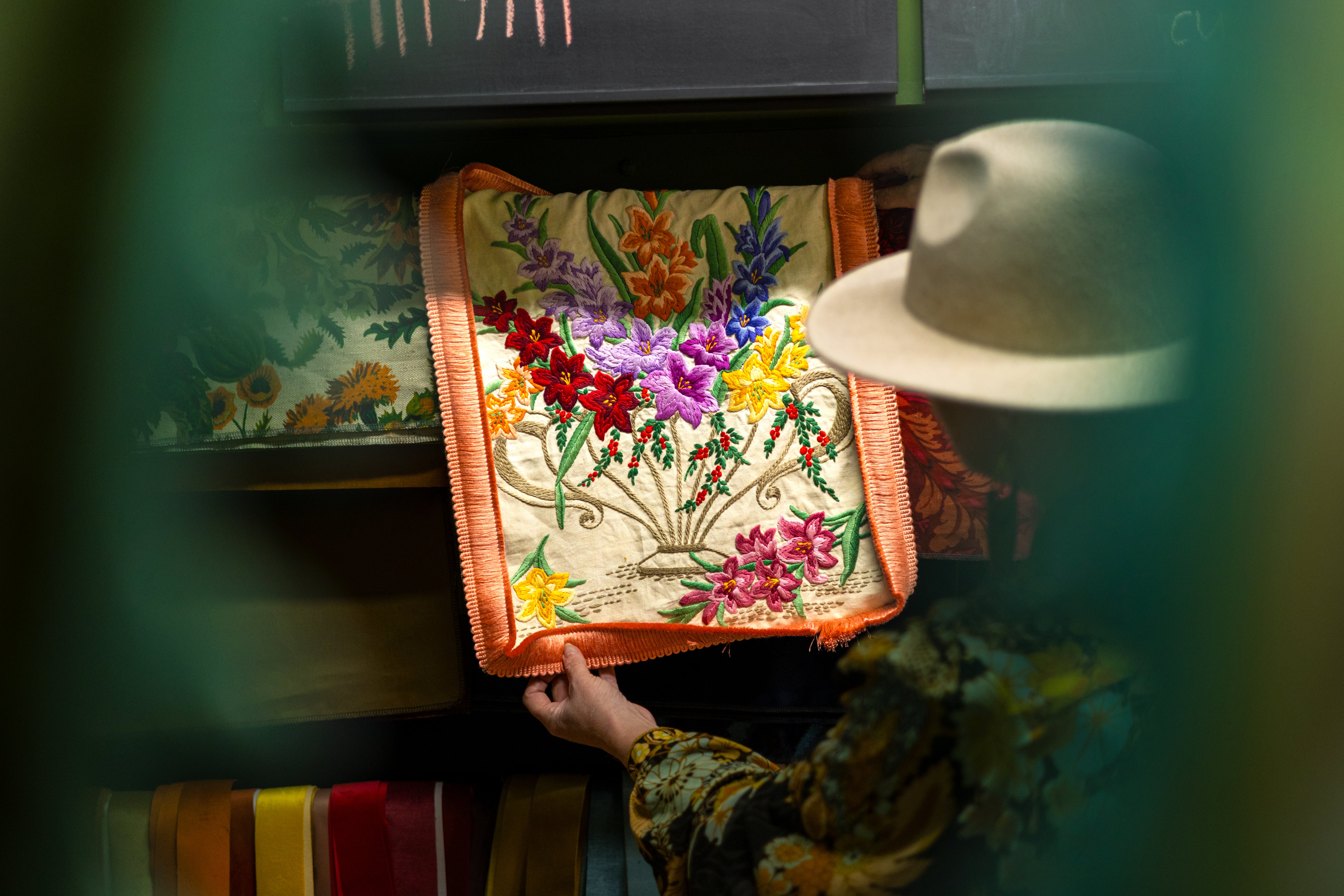 A person wearing a light-colored hat holds up a square embroidered cushion cover featuring a colorful floral bouquet design with orange borders, shown in a dimly lit interior setting.