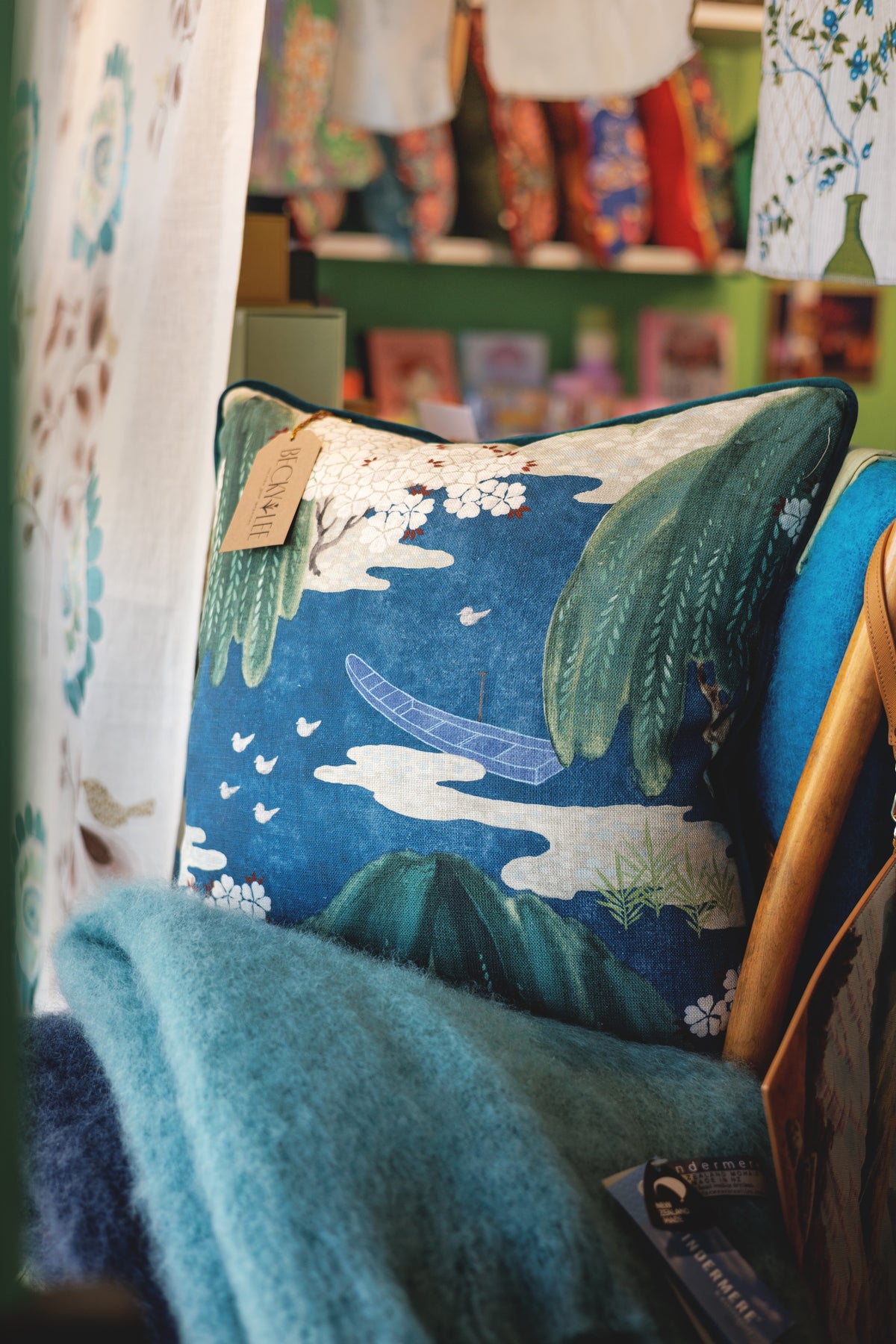 A square decorative throw pillow with a blue and green landscape print featuring clouds, birds, and a boat, styled on a teal blanket in a shop setting.
