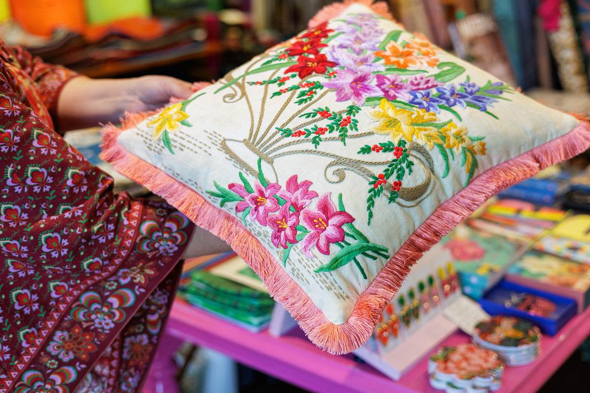 A square decorative throw pillow with vibrant floral embroidery and coral fringe trim, held by a person in a patterned dress in what appears to be a craft or retail shop.