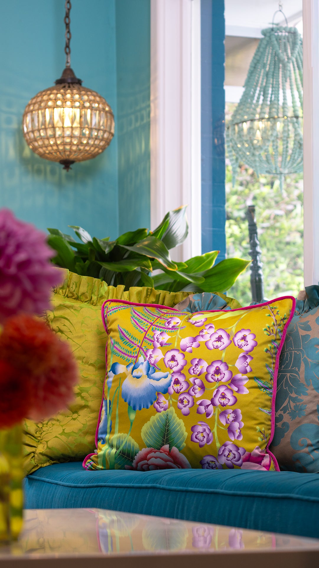 Colorful living room with a yellow floral cushion on a blue couch, decorative plants, and hanging lights.