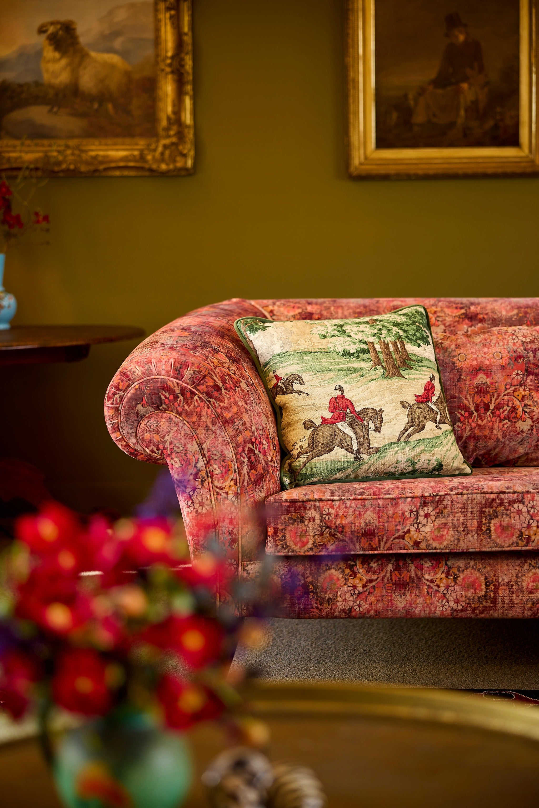 Photo of a luxurious room with vintage artworks, a rolled arm sofa upholstered in an antique damask style pattern and a cushion depicting horses, hounds and riders on a traditional hunt.