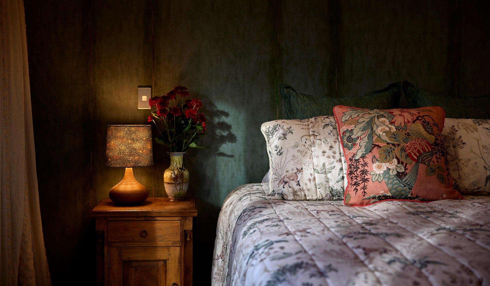 Photo of a moody bedroom setting with murky turquoise wallpaper, timber bedsides and a salmon coloured floral linen cushion