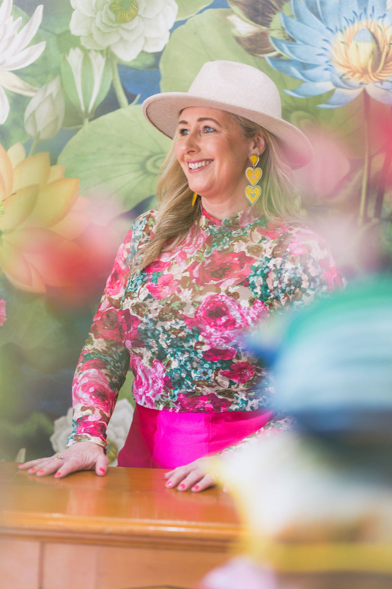 Woman in floral blouse and pink skirt standing in front of a colorful floral background