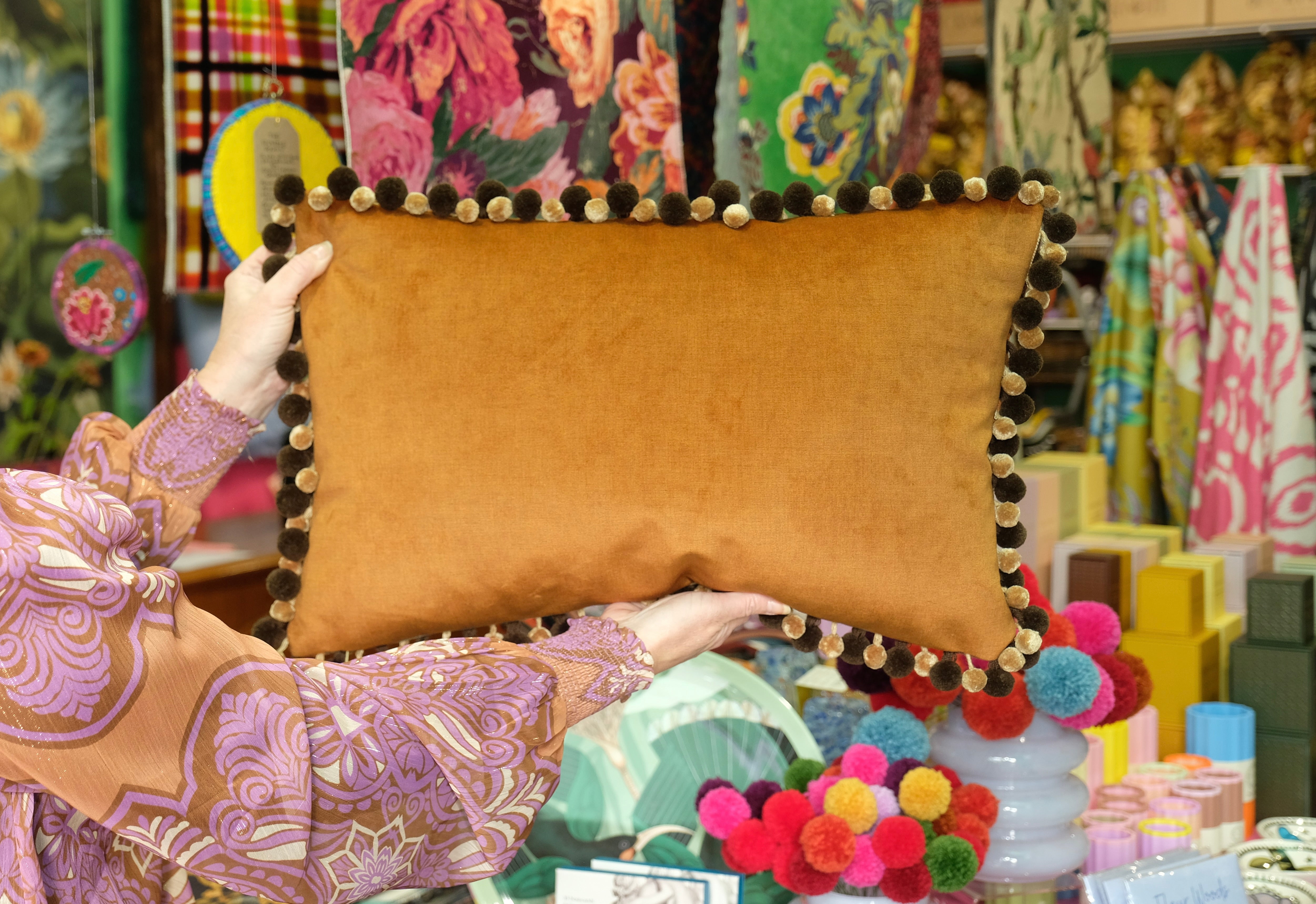Person holding a brown pillow with pom-pom trim in a colorful store setting