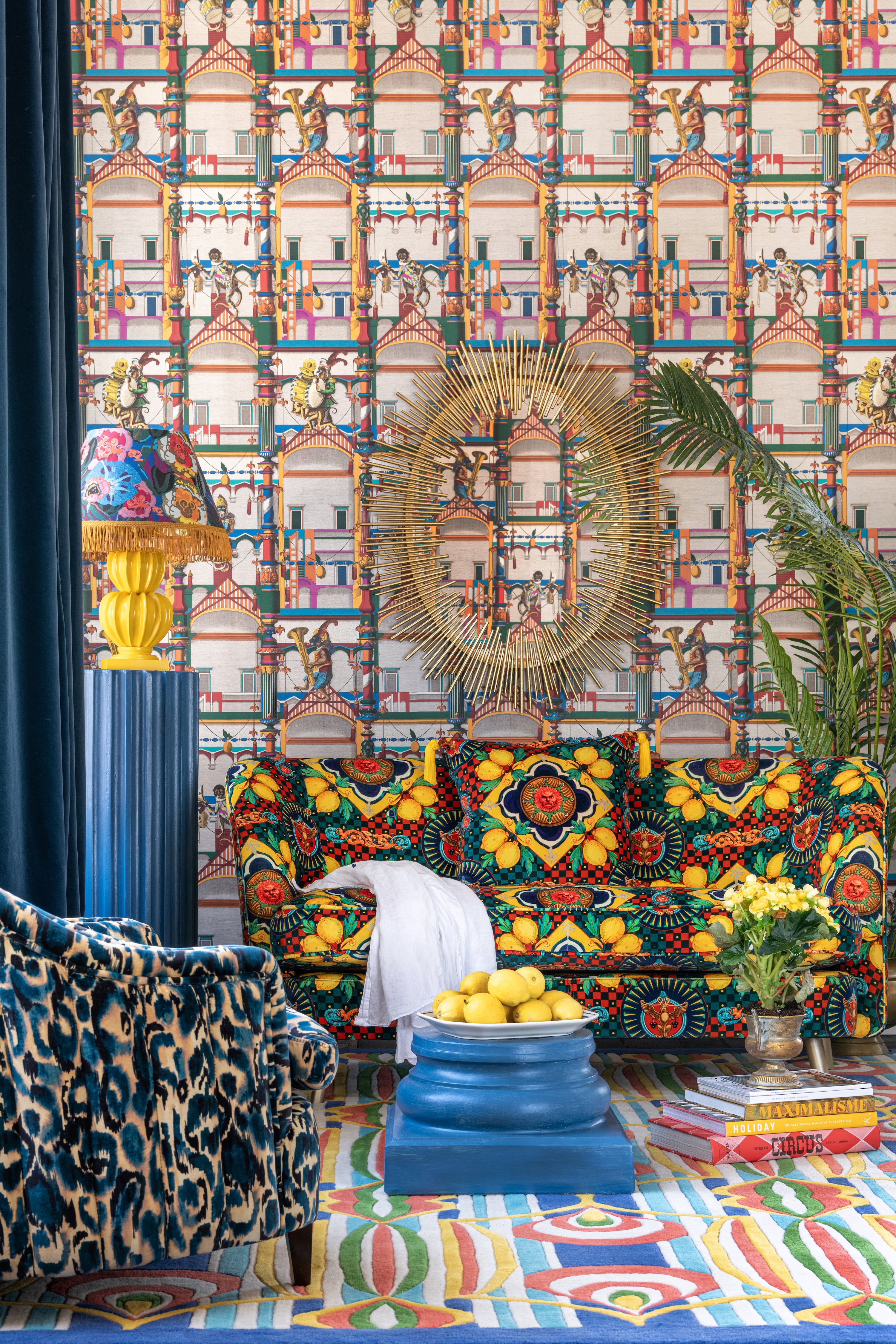 Luxe living space with ornate architectural EL CIRCO wallpaper, a bold patterned sofa, a blue leopard-print chair, and a striking gold sunburst mirror.