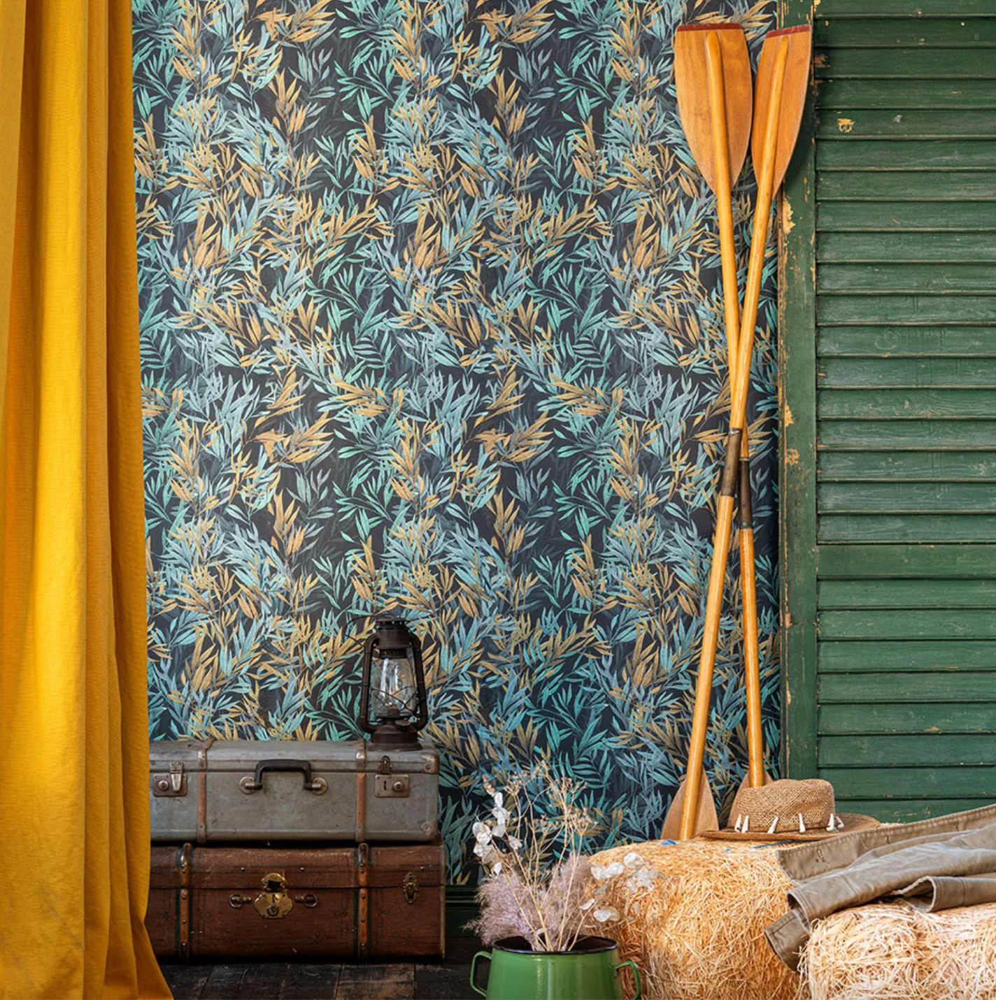 A rustic setting with leafy-patterned wallpaper in blue and gold tones, wooden oars, vintage trunks, a lantern, and hay bales with cozy decor accents
