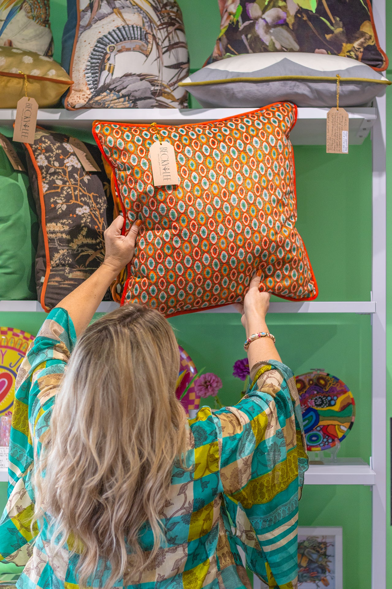 Woman holding an orange patterned pillow against a green wall with shelves displaying various pillows.
