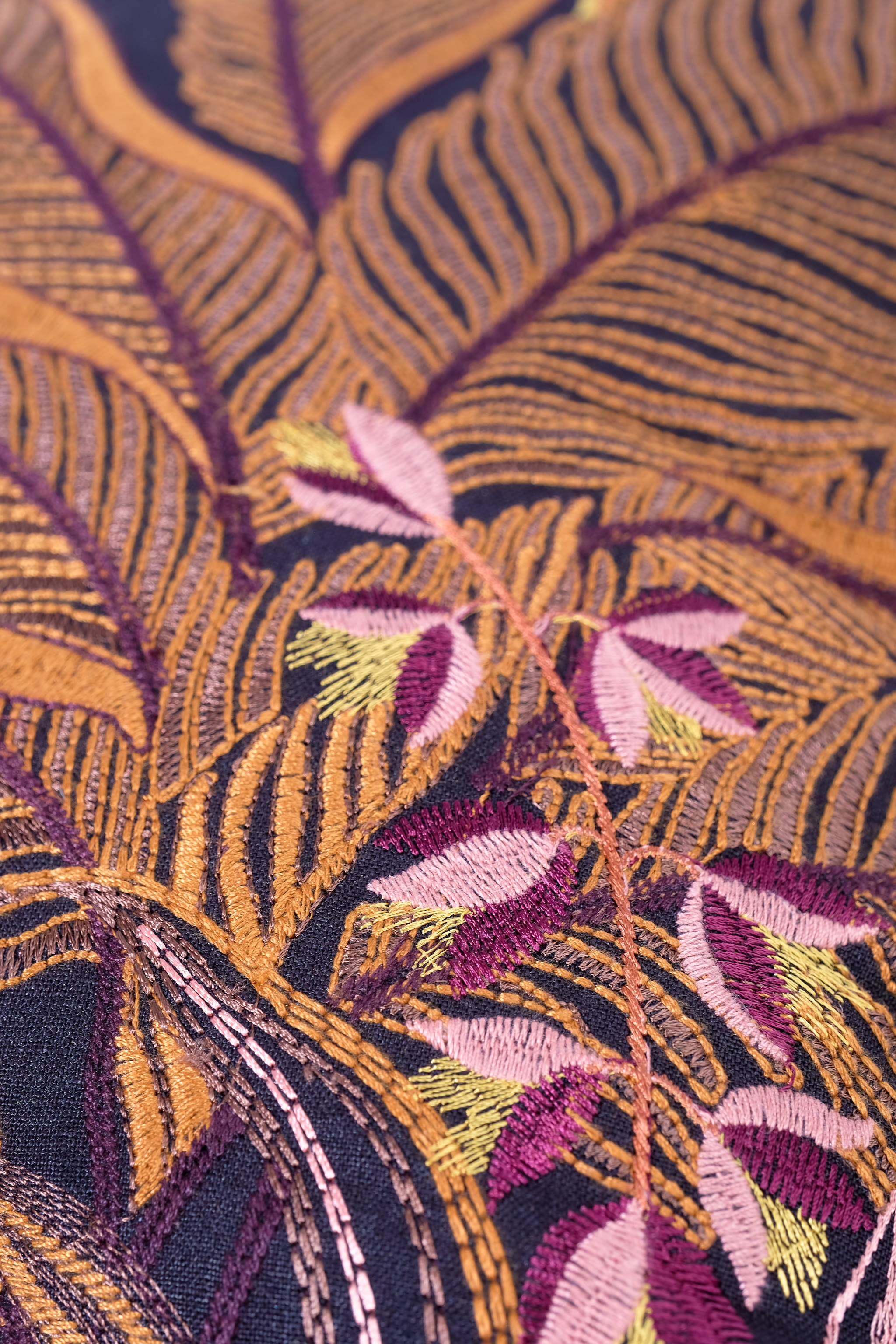 Close-up of an embroidered fabric with a tropical leaf pattern in gold, purple and pink on a dark background.