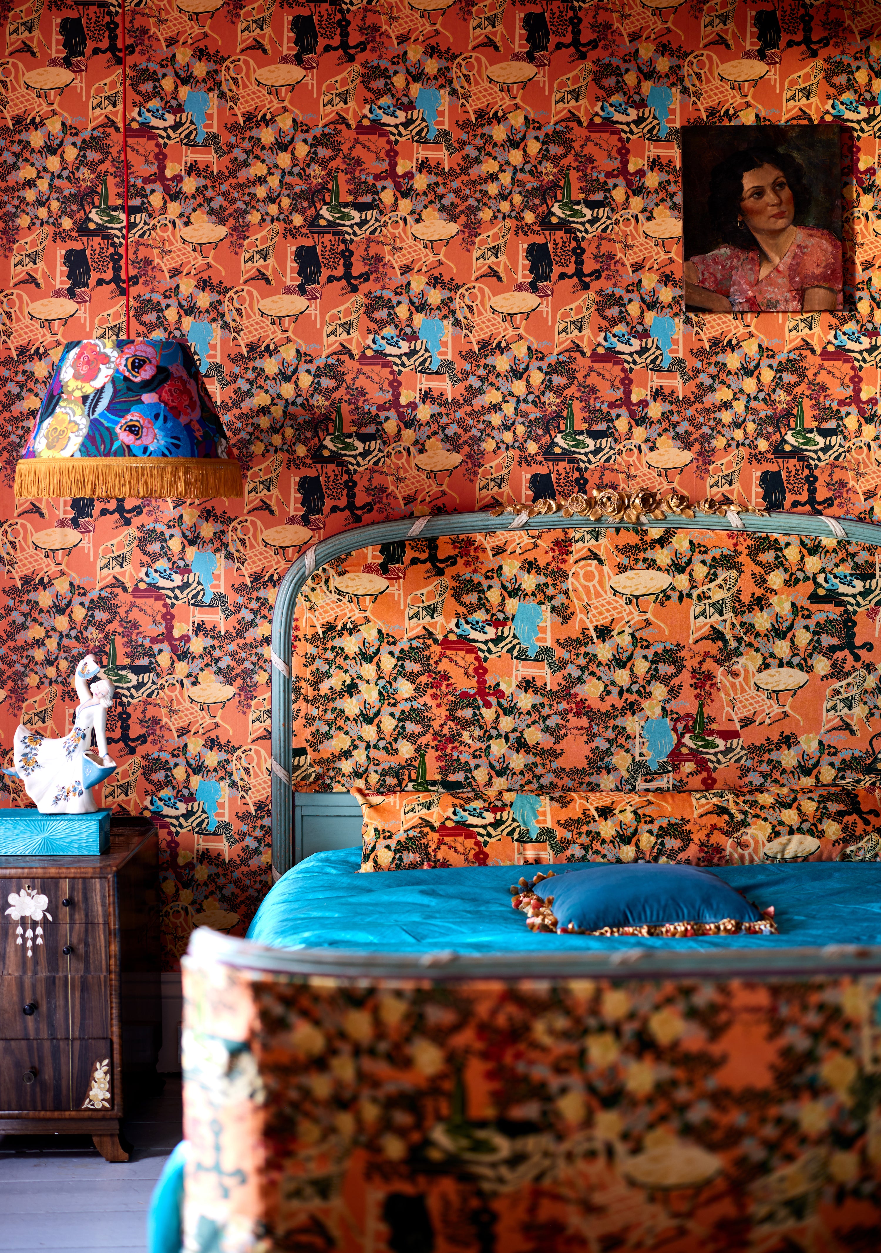Bedroom with LA BRASSERIA Salmon wallpaper featuring vintage café scenes, a matching upholstered bed, blue bedding, a fringed lamp, and antique decor