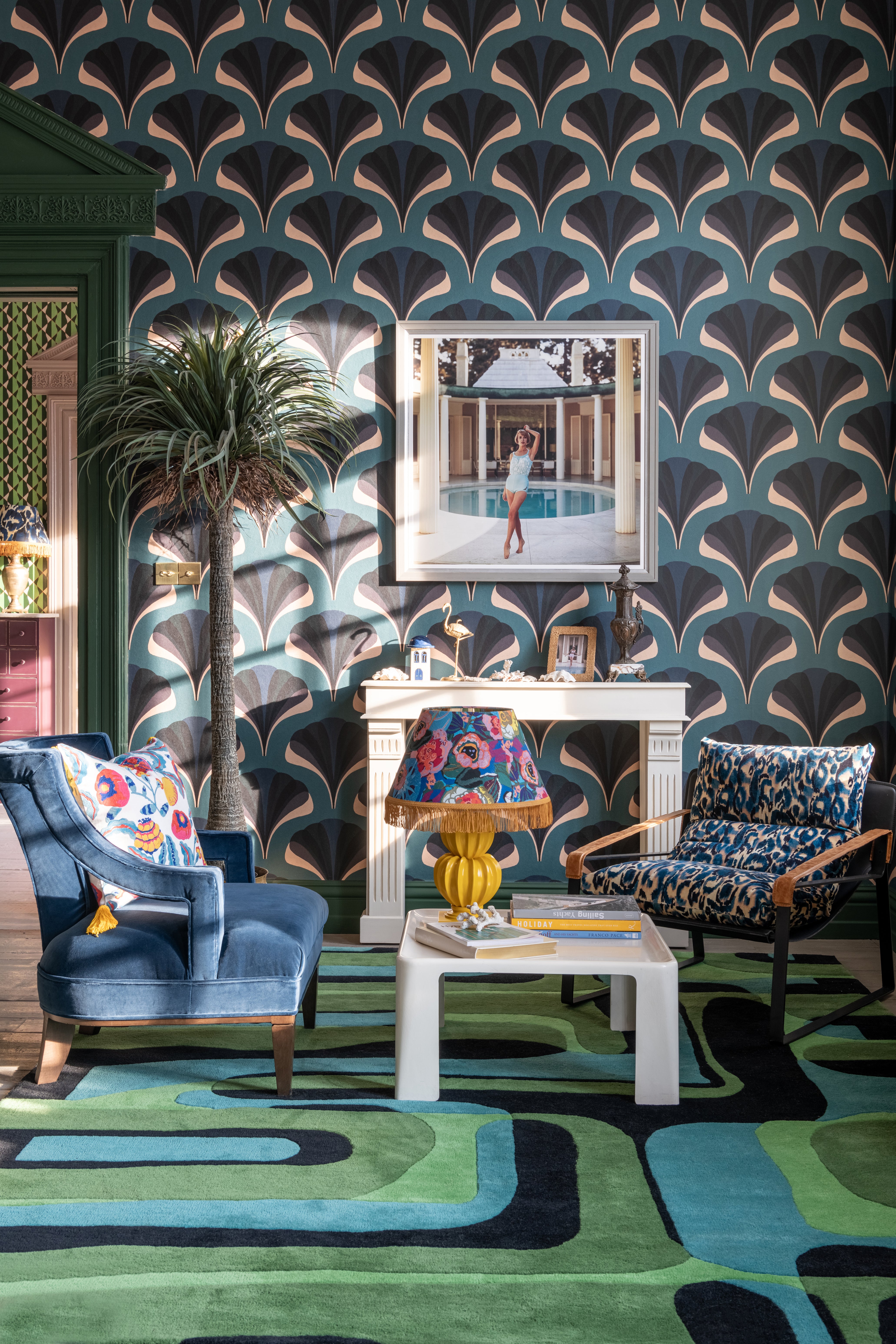 Elegant living room with MEZZELUNE Kale wallpaper, a bold green and blue rug, velvet armchairs, a fringed lamp, and a stylish faux fireplace.




