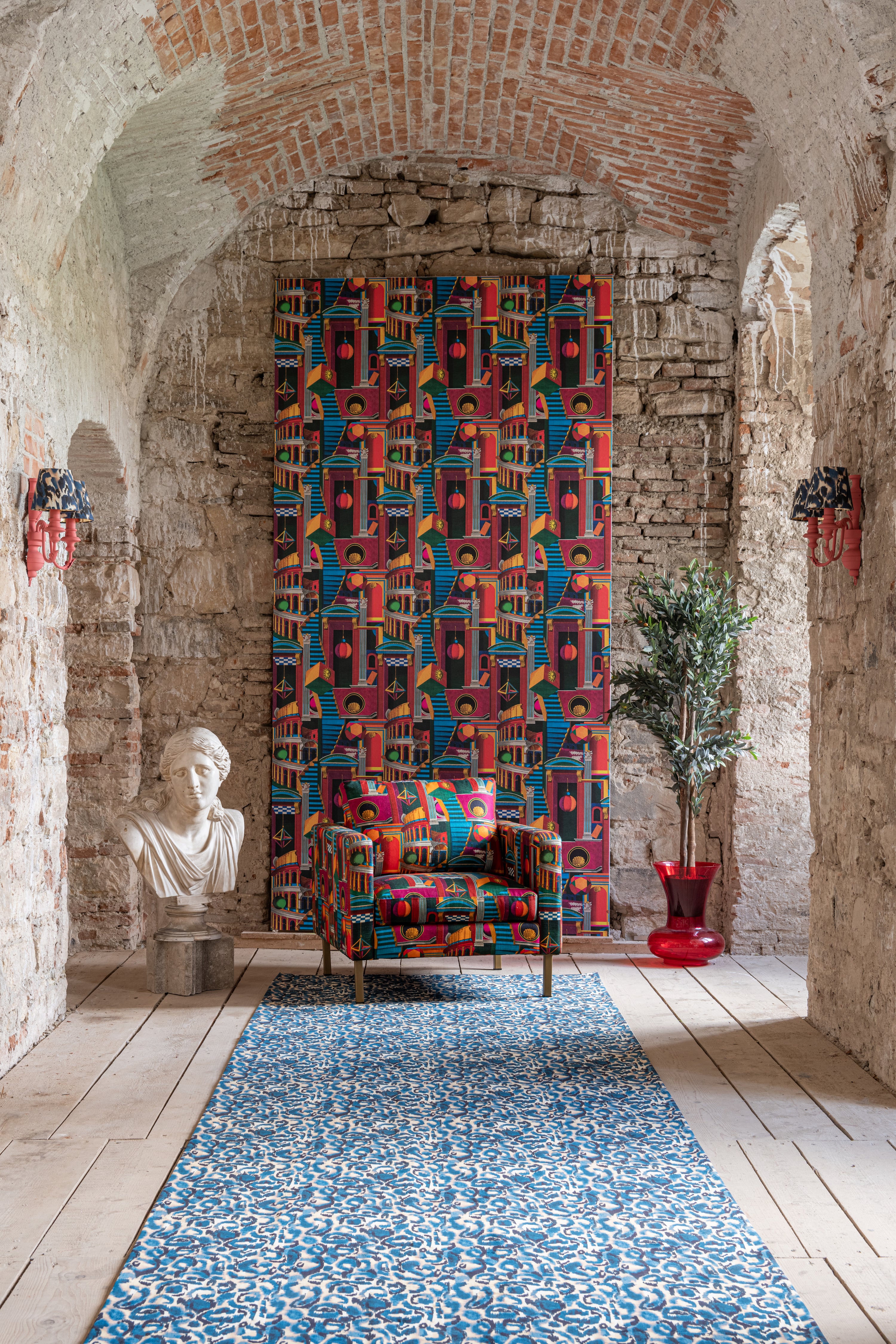 Eclectic space with exposed brick, a blue patterned rug, and a bust statue. PIAZZA DEGLI ARLECCHINI wallpaper is hanging, on a panel in the centre back with a chair in front with a matching pattern 