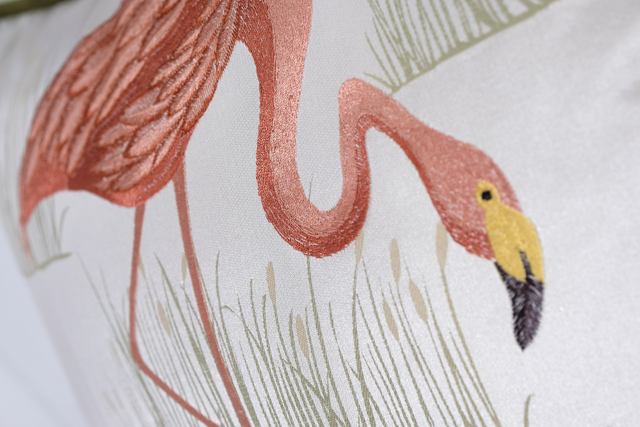 Close-up of a decorative textile featuring an embroidered pink flamingo with a yellow beak and muted green grass on a light background.