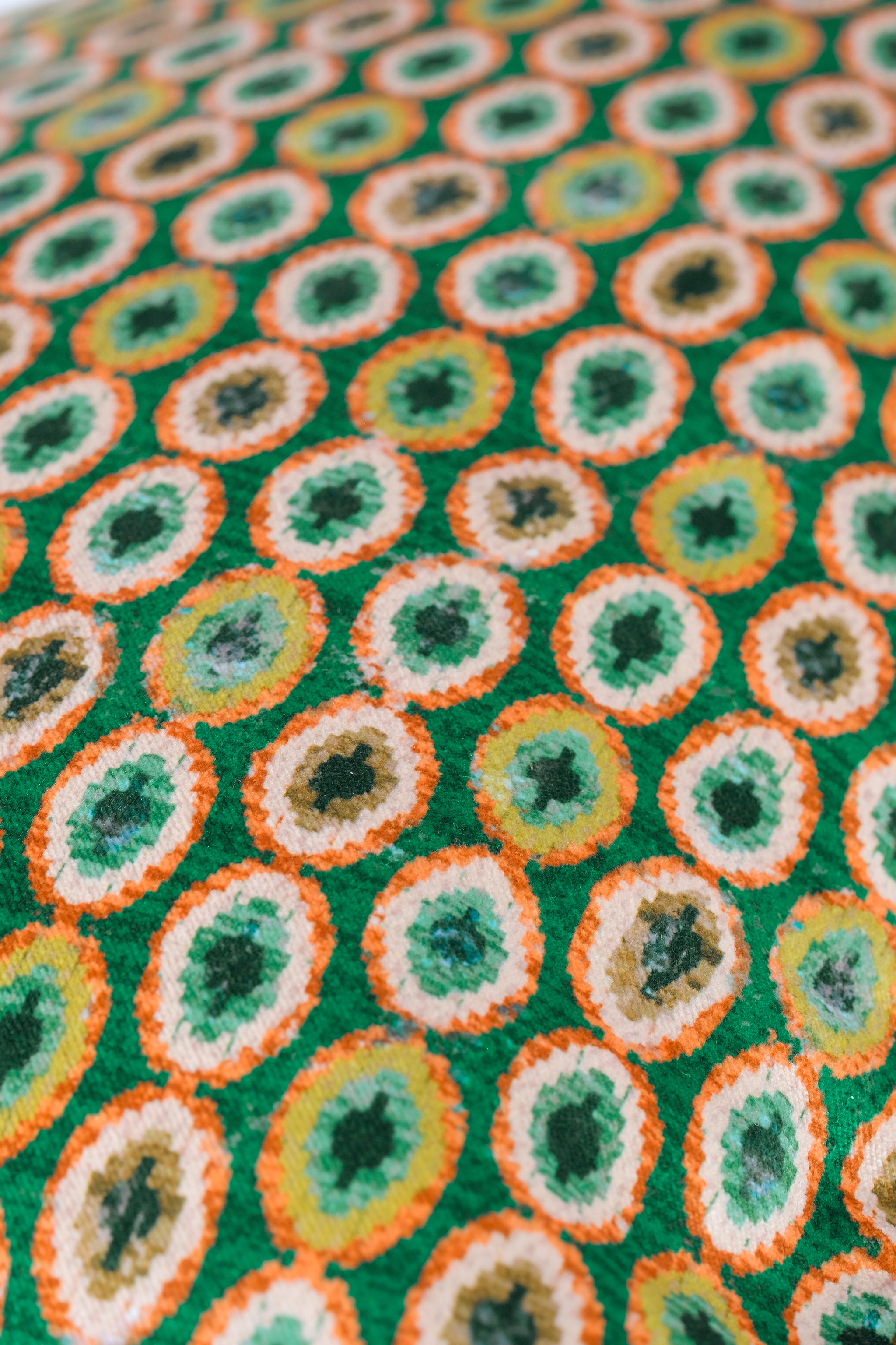 A close-up of a green textured fabric with a repeating circular pattern in orange, white, yellow, and dark green.