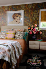 Bedroom with floral wallpaper, bed with colorful pillows, and a vase of flowers on a nightstand.