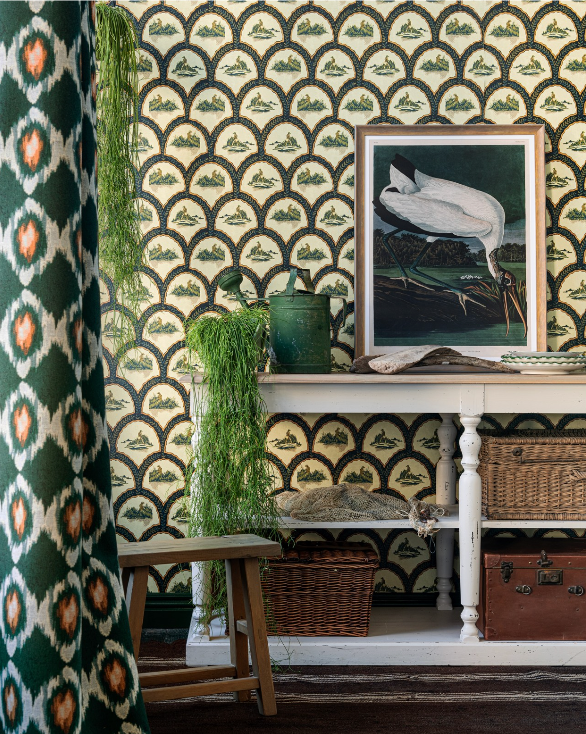 Vintage-inspired wallpaper with arched landscape motifs in cream and green. A white wooden table is decorated with plants, baskets, and a framed bird print.