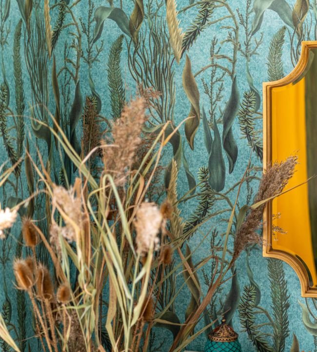 A close-up of an aquatic-themed wallpaper in deep blue-green hues, featuring underwater plants. Dried grasses and a gold mirror add warmth and texture.